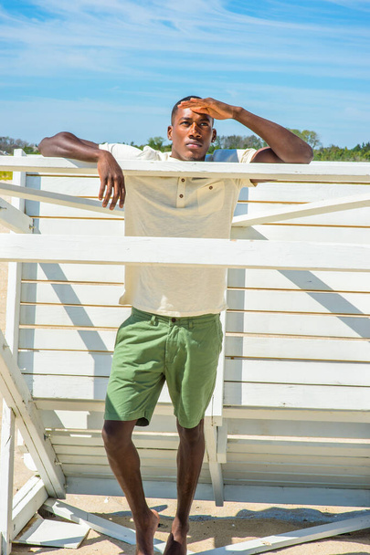 Одна рука поднимается на лоб, молодой красивый черный парень стоит у белого деревянного строения на пляже, глядя вперед - Фото, изображение