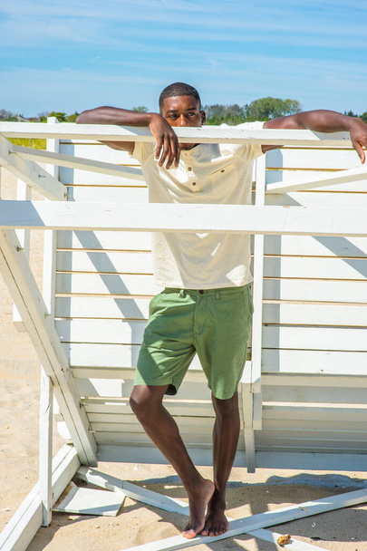 Beide Arme auf einen Stock gestützt, steht ein junger, gutaussehender schwarzer Mann an einer weißen Holzkonstruktion am Strand und schaut, entspannt und nachdenklich. - Foto, Bild