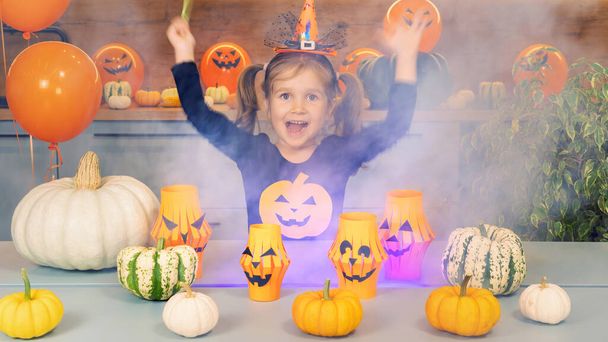 Die kleine Halloween-Hexe zaubert mit einem Zauberstab Kürbisse. Glückliches Kind lächelt und spielt Halloween. Ein spitzbübisches Mädchen führt als Zauberin verkleidet Zaubertricks mit buntem Rauch auf. - Foto, Bild