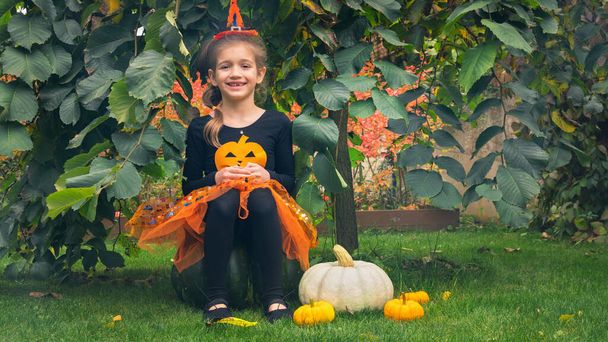 Little witch girl sitting on a pumpkin for Halloween. A girl in a witch costume hides under the umbrella elm tree and smiles. Children's trick or treat carnival concept. - Photo, Image