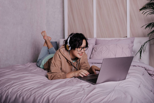 Egy kaukázusi tinilány fekszik az ágyon fejhallgatóval és laptoppal. - Fotó, kép