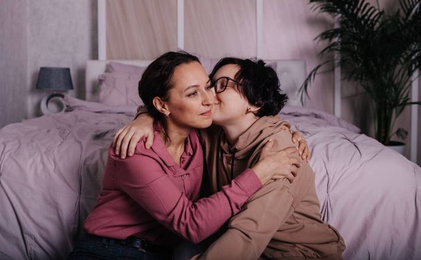 een Kaukasische tiener dochter kust een brunette moeder in een kamer in de buurt van het bed - Foto, afbeelding