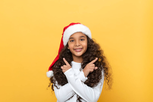 Waist up portrait of a dark skinned young girl with long curly hair in a Santa Claus hat against yellow background - Zdjęcie, obraz