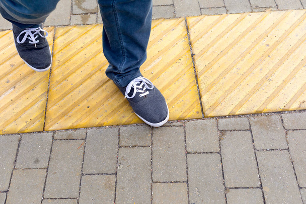 Męskie stopy na chodniku z jasnożółtą, dotykową powłoką dla osób o słabym wzroku. Infrastruktura, technologie wspomagające dla osób niepełnosprawnych - Zdjęcie, obraz