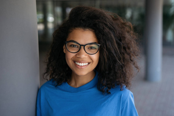 Porträt einer jungen lächelnden Afroamerikanerin mit stylischer Brille, die auf der Straße steht. Glücklich selbstbewusste Studentin blickt auf Uni-Campus in die Kamera - Foto, Bild