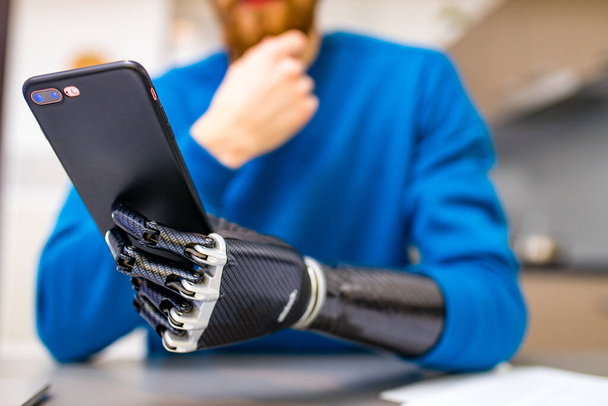young man with bionic hand hold smartphone while sitting indoors home kitchen background - Foto, immagini