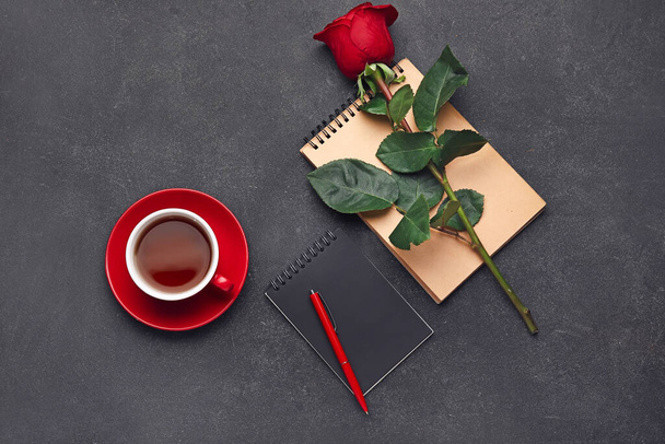 Piękna czerwona róża, zeszyty i filiżanka herbaty na ciemnym tle - Zdjęcie, obraz