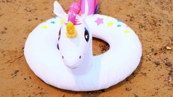Plajda kumsalda beyaz bir tek boynuzlu at şeklinde şişirilebilir bir yüzük. Denize açılmak için fantezi yüzme ringi. Yaz tatili konsepti, güneşli gün ve sıcak hava. - Video, Çekim