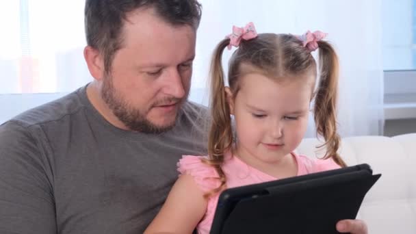 vicces kislány 3 éves és az apja együtt tanul egy táblagépen, és néz egy leckét online otthon. Apa segít a lányának a tanulásban. Online iskolai távoktatási koncepció. - Felvétel, videó