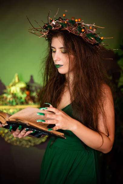 Witch with disheveled hair and long nails with a wreath of dry branches on her head, dressed in a green dress, posing with a black book with a pentagram on the cover standing  next to a cauldron. - Foto, afbeelding