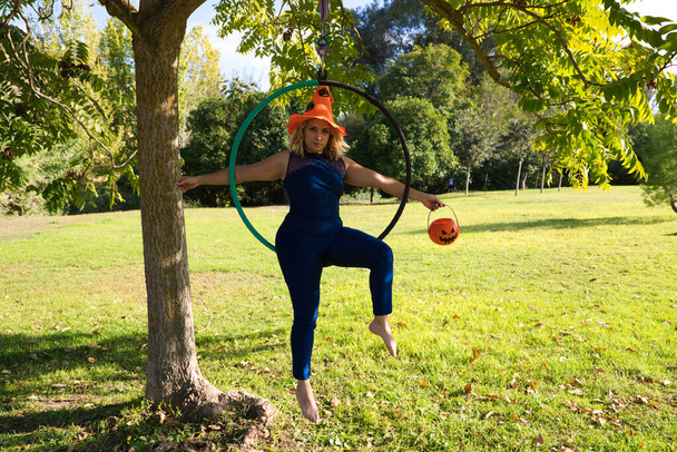 Взрослая акробатическая спортсменка в парке занимается спортом. У женщины есть оранжевая шляпа на Хэллоуин и тыква в руке во время акробатики. Здоровье и спорт. - Фото, изображение