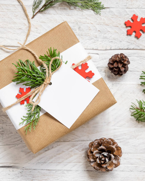 Weihnachten verpacktes Geschenk mit quadratischem Papier Geschenkanhänger auf einem weißen Tisch mit Tannenzweigen und Dekorationen Draufsicht. Rustikale Winterkomposition mit leerem Geschenkanhänger Mockup, Kopierraum - Foto, Bild