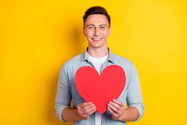 Portrait of attractive cheerful guy holding in hands big heart card isolated over bright yellow color background - Foto, Imagen