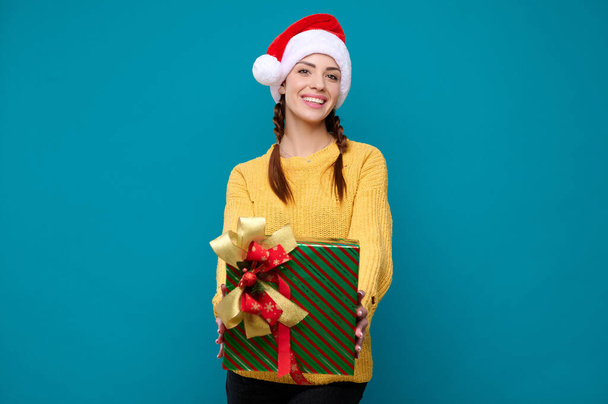 Schöne junge lächelnde Frau mit Weihnachtsmann-Hut, die ein Neujahrsgeschenk auf blauem Hintergrund hält. Neujahrskonzept - Foto, Bild