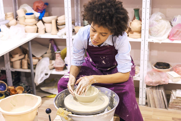Creative afro american young woman molding clay on pottery wheel. Workshop in ceramic studio. Female artist.  - Photo, Image
