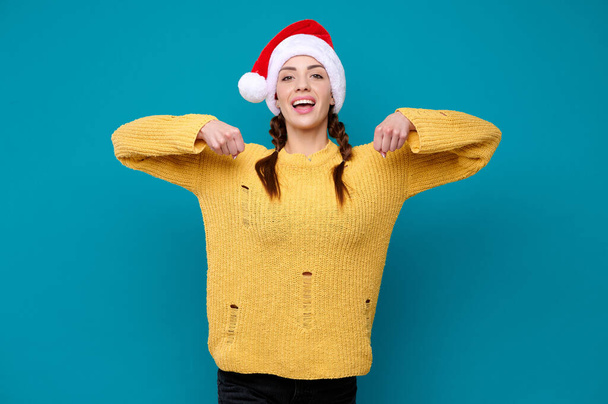 Young smiling woman in yellow sweater and santa hat holding an invisible banner and blue isolated background. Product advertisement. New Year concept - Foto, Imagem