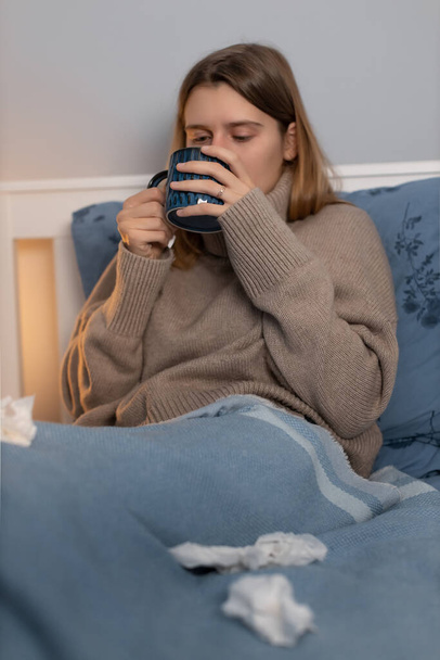 Cute woman sitting in bed and drinking hot beverage. Girl feels fever, headache and weakness. Female got sick with flu, cold, angina or sore throat. Treatment at home. Selective focus on hands. - Photo, Image