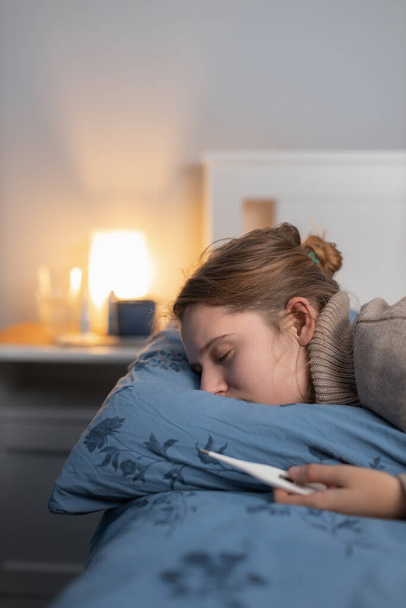 Mujer caucásica demacrada en suéter beige se durmió en la cama con termómetro en la mano. Mujer agotada por la enfermedad. Niña que sufre de resfriado y gripe. Concepto de salud. Copiar espacio. - Foto, imagen