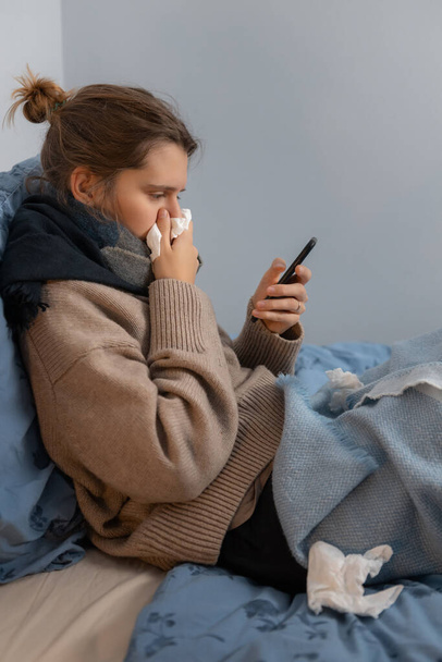 Disease cute caucasian woman in beige sweater and black scarf blowing nose into paper napkin. Girl sitting in bed and searching medicines delivery in internet. Online purchase of medicines. - Photo, Image