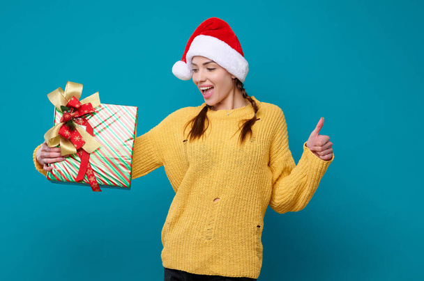 Happy surprised young woman in santa claus hat holding new year gift laughing and showing thumb up gesture on blue isolated background - Photo, Image
