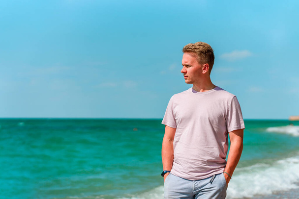 Egy fiatal szőke férfi sétál a parton az azúrkék óceán vagy a tenger közelében. A boldog nyári vakáció koncepciója - Fotó, kép