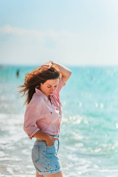 A charming young woman with long hair and wearing a shirt walks along the beach on a sunny day along the azure sea or ocean. The concept of summer holidays - Photo, image