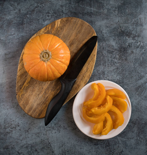 A small ripe pumpkin on a wooden chopping board next to a knife and sliced pumpkin on a white plate on a dark background. Traditional autumn food. Top view - Valokuva, kuva