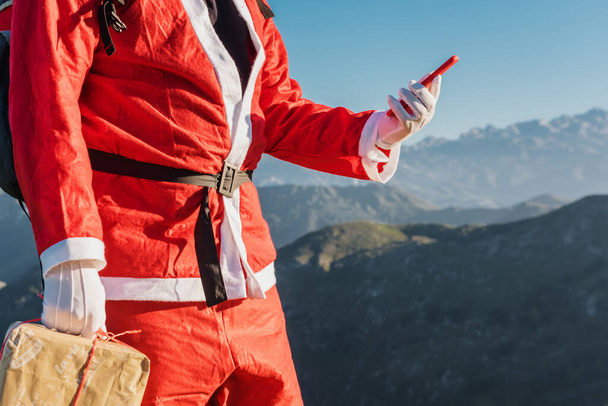 De kerstman gebruikt een app op zijn smartphone om geschenken te bezorgen op afgelegen berglocaties. Kerstconcept - Foto, afbeelding
