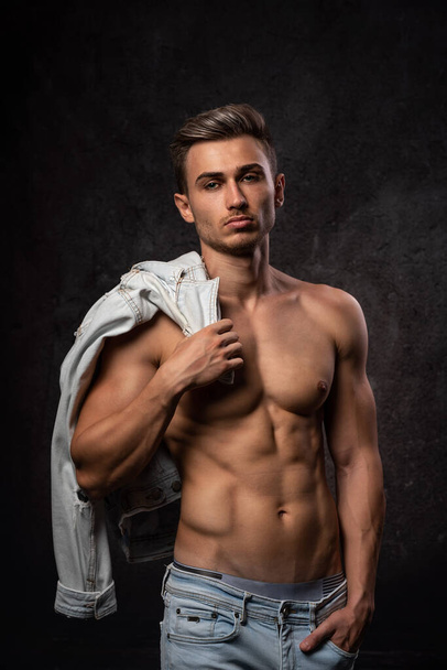 European athletic young man, posing without outerwear, in sports physical form. On a dark textured background. Jacket on the shoulder. - Foto, Imagem