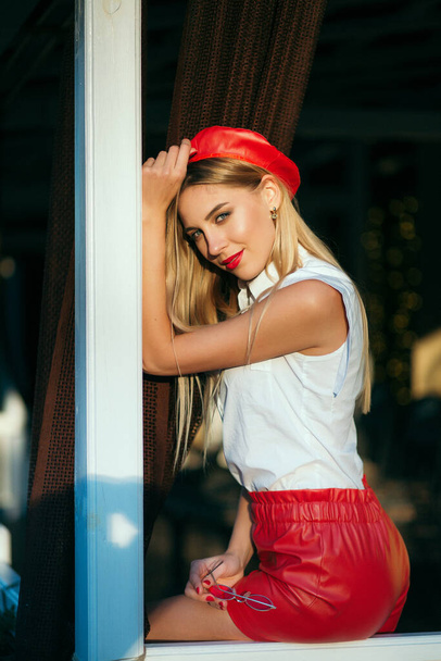 Portrait of young blonde female in red beret and leather red shorts posing outdoor. Bright make up. Red glasses and scarf. - Photo, Image