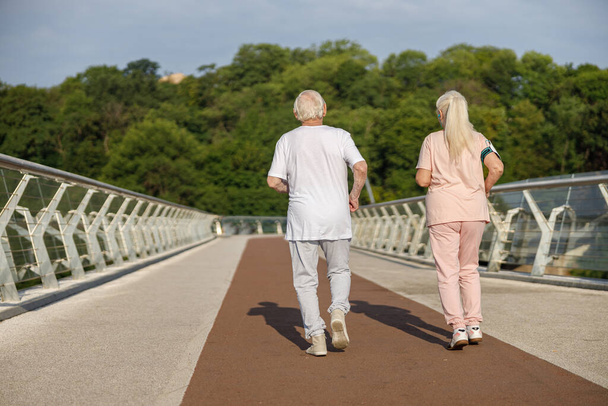 Sportive senior man and woman in tracksuits run together along footbridge - Photo, Image