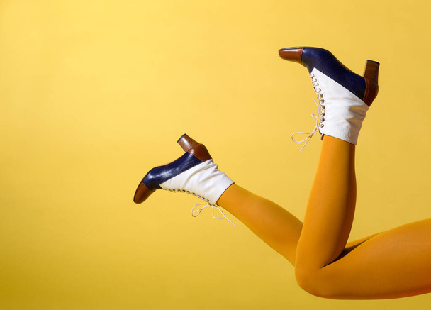 Side view of crop anonymous fashionable slim female model wearing stylish retro tricolor leather boots and mustard colored pantyhose lying on yellow background - Photo, Image