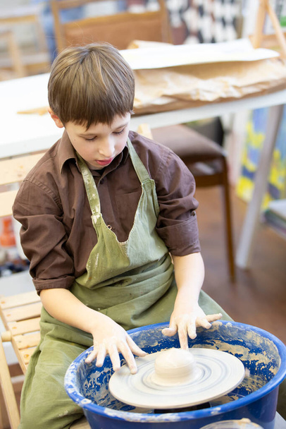 Ένα μικρό αγόρι φτιάχνει ένα πήλινο προϊόν στον τροχό ενός αγγειοπλάστη.. - Φωτογραφία, εικόνα