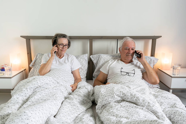senior aging married couple man male and female woman resting together in bed at home apartment using call smartphone - Valokuva, kuva
