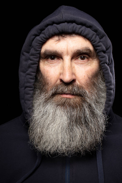 An elderly man with a gray beard in a hood on a black background. - 写真・画像