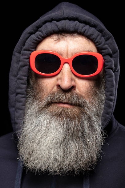 An elderly man with a gray beard in a hood and red glasses on a black background. A man hides something. - Photo, Image