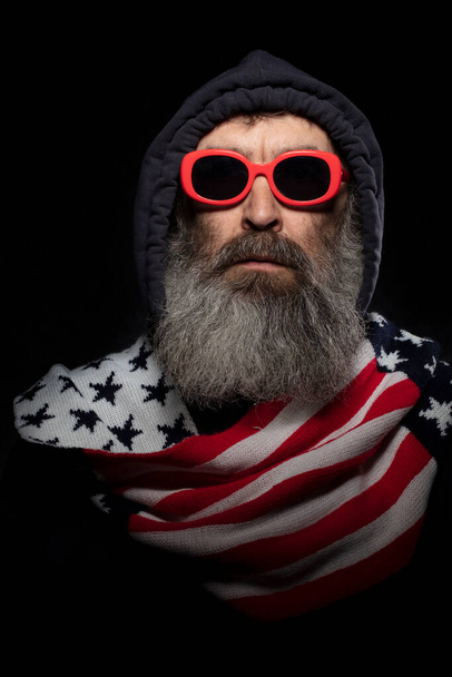Літній чоловік з сірою бородою в капюшоні і шарф з візерунком американського прапора на чорному тлі.. - Фото, зображення