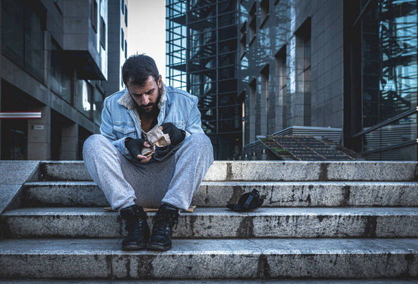 Young hungry dirty depressed homeless man sitting on the stairs of the building on sidewalk and try to eat food scraps from paper bag that he find in the garbage can, homelessness social concept - 写真・画像