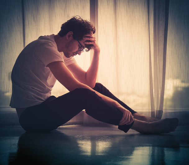 Homem caucasiano desesperado sentado sozinho à janela em silhueta com tristeza no chão. - Foto, Imagem