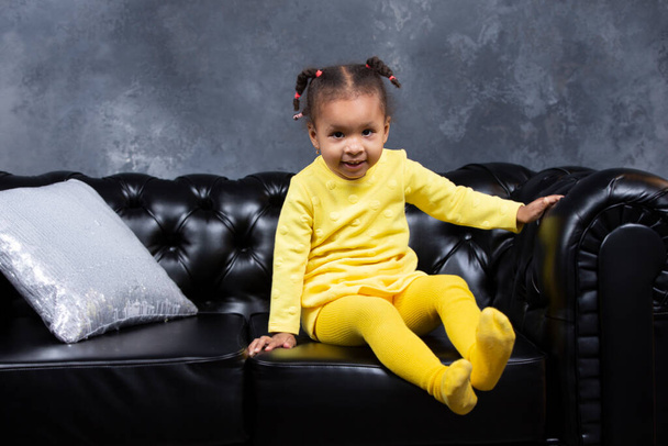 Niña afroamericana se sienta en un sofá de cuero negro. - Foto, imagen