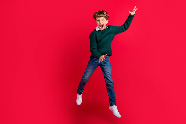 Photo of funny charming school boy wear knitted pullover smiling jumping high isolated red color background - Foto, afbeelding
