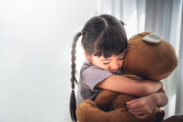 Asyalı kızın bulanık görüntüsü 6 yaşında, büyük kahverengi bir ayıya sarılıyor, yalnızlık ve üzüntü içinde, beyaz sırtında, çocuklara ve aile kavramına. - Fotoğraf, Görsel
