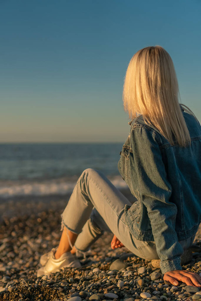 Die junge blonde Frau sitzt an einem Kieselstrand am Meer und betrachtet den Sonnenuntergang, ein Foto von ihrem Rücken. Konzept des Reisens, des Genusses, der Einsamkeit. Vertikales Foto - Foto, Bild