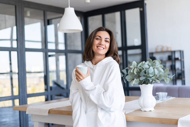 Jovem mulher em um capuz branco quente em casa na cozinha, começa o dia com uma xícara de café, posando, de bom humor, sorrindo - Foto, Imagem