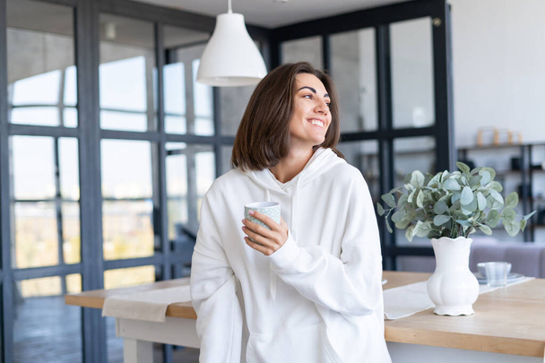 Jonge vrouw in een warme witte hoodie thuis in de keuken, begint haar dag met een kopje koffie, poseren, in een goede bui, glimlachen - Foto, afbeelding