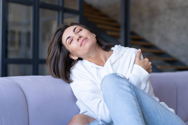 Een jonge vrouw thuis in een witte hoodie op de bank zit vredig, geniet van eenzaamheid, comfort, alleen, in een rustige stemming - Foto, afbeelding
