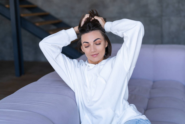 A young woman at home in a white hoodie on the couch sits peacefully, enjoys loneliness, comfort, alone, in a calm mood - Photo, Image