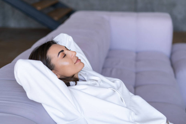 Junge Frau zu Hause im weißen Kapuzenpulli auf der Couch sitzt gelassen mit geschlossenen Augen, entspannt, ruht, meditiert - Foto, Bild