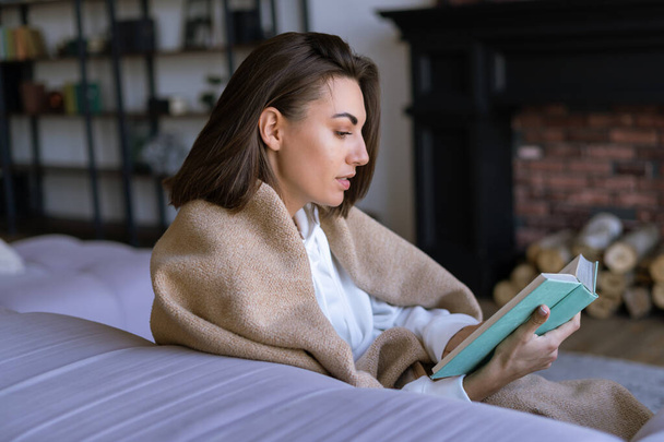 Młoda kobieta w białym kapturze na kanapie owinęła się ciepłym kocem, czytała książkę, powieść, zimowy wieczór - Zdjęcie, obraz