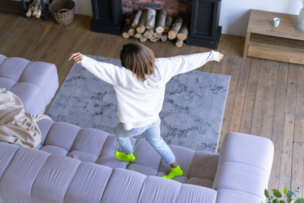 Young woman at home in a white hoodie and jeans jumping on the sofa, dancing, having fun - Fotoğraf, Görsel